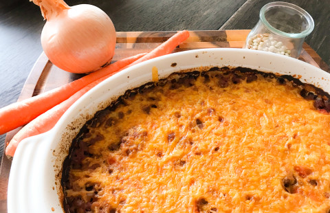 casserole dish of baked lentils with melted cheddar cheese on top with fresh onion and carrots in the background